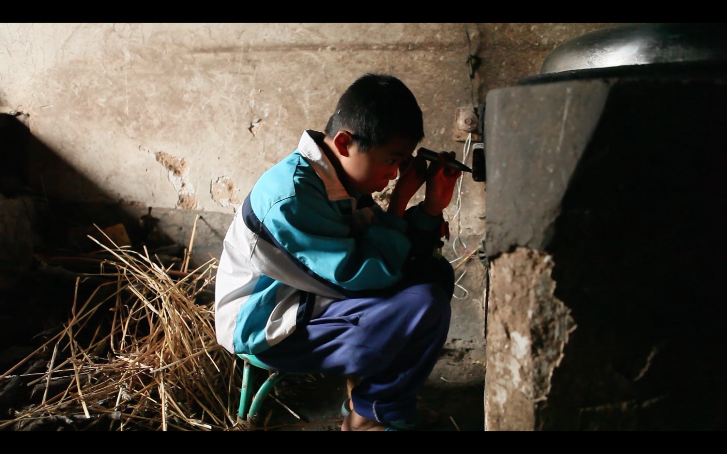 A young Chinese boy, maybe ten years old, sits on a low stool, working intensively on an electrical device attached to an old decaying structure from the film Under the Same Sky.